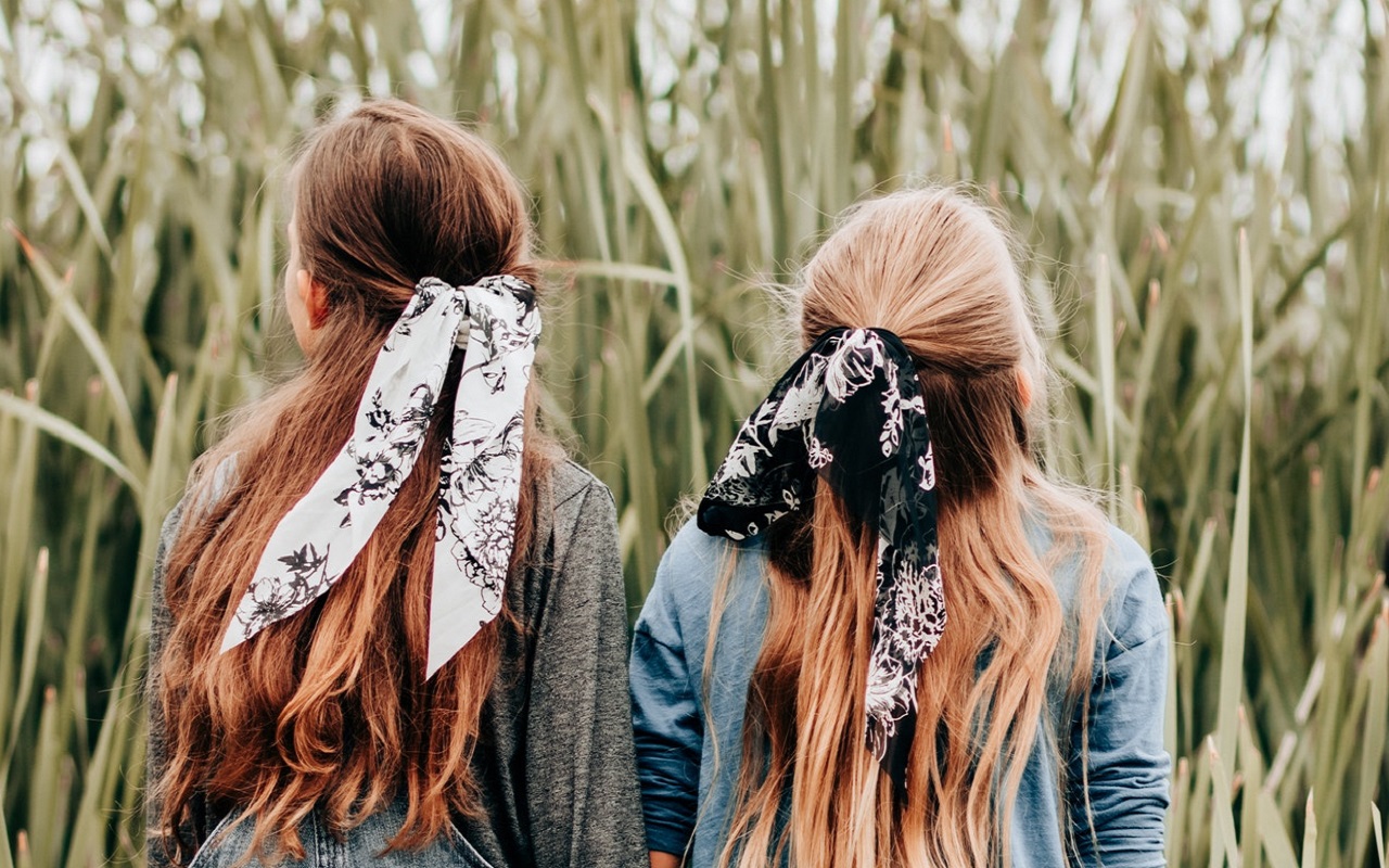 Odwrócone tyłem dwie dziewczynki z długimi włosami mają związane włosy kolorowymi wstążkami. Przed nimi wysokie trawy i zboża