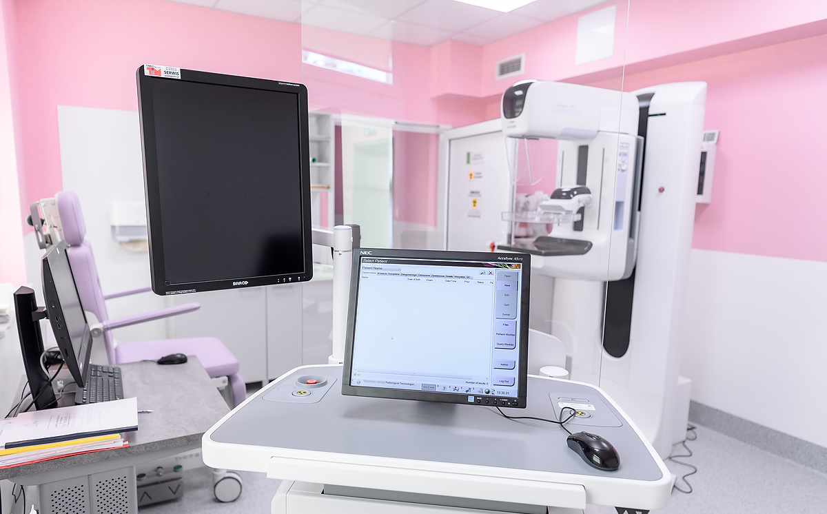 Pomieszczenie o różowych ścianach, które jest pracownią mammograficzną. Na pierwszym planie widać monitory, służące do odczytu wyników badania, za nimi stoi urządzenie do wykonywania badania piersi - mammograf