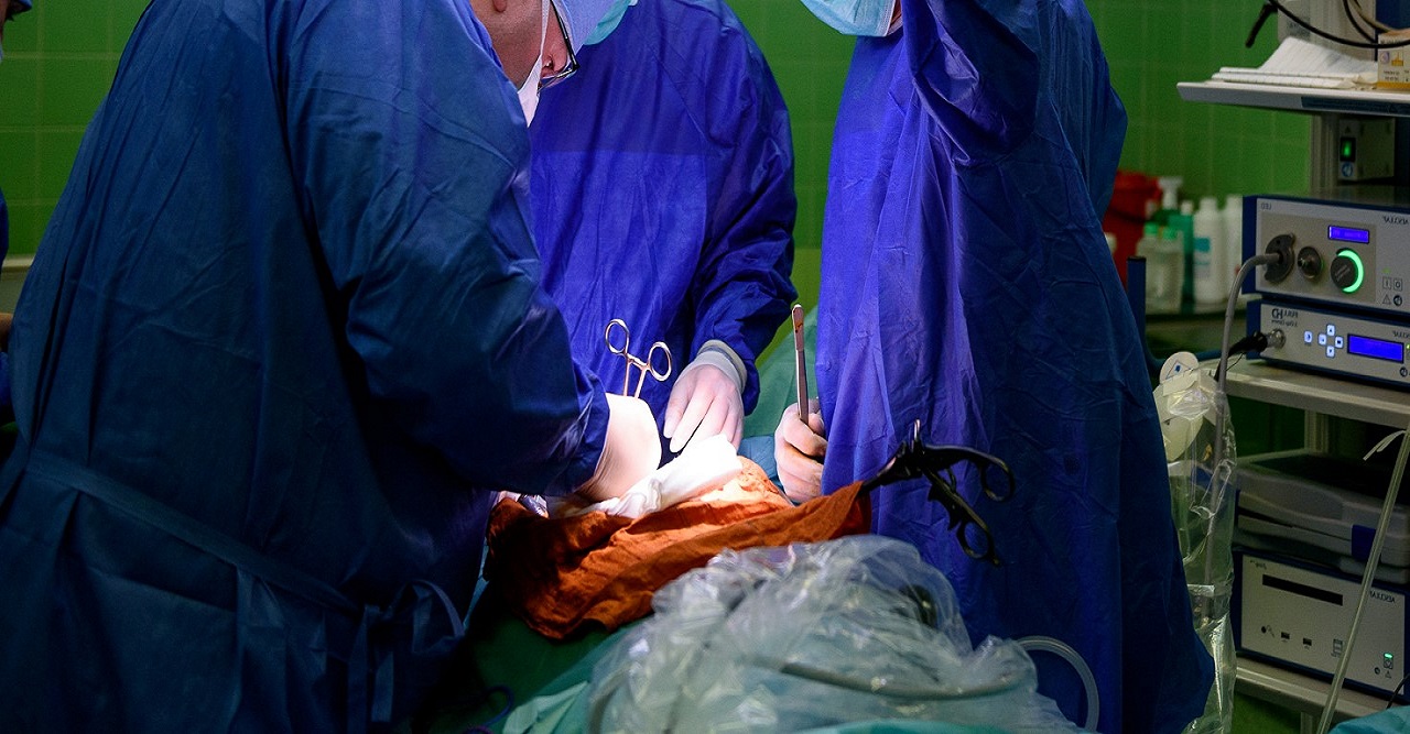 Sala operacyjna, na pierwszym planie widoczny personel medyczny prowadzący operację: chirurg oraz osoby asystujące. Na drugim planie widoczna jest aparatura medyczna