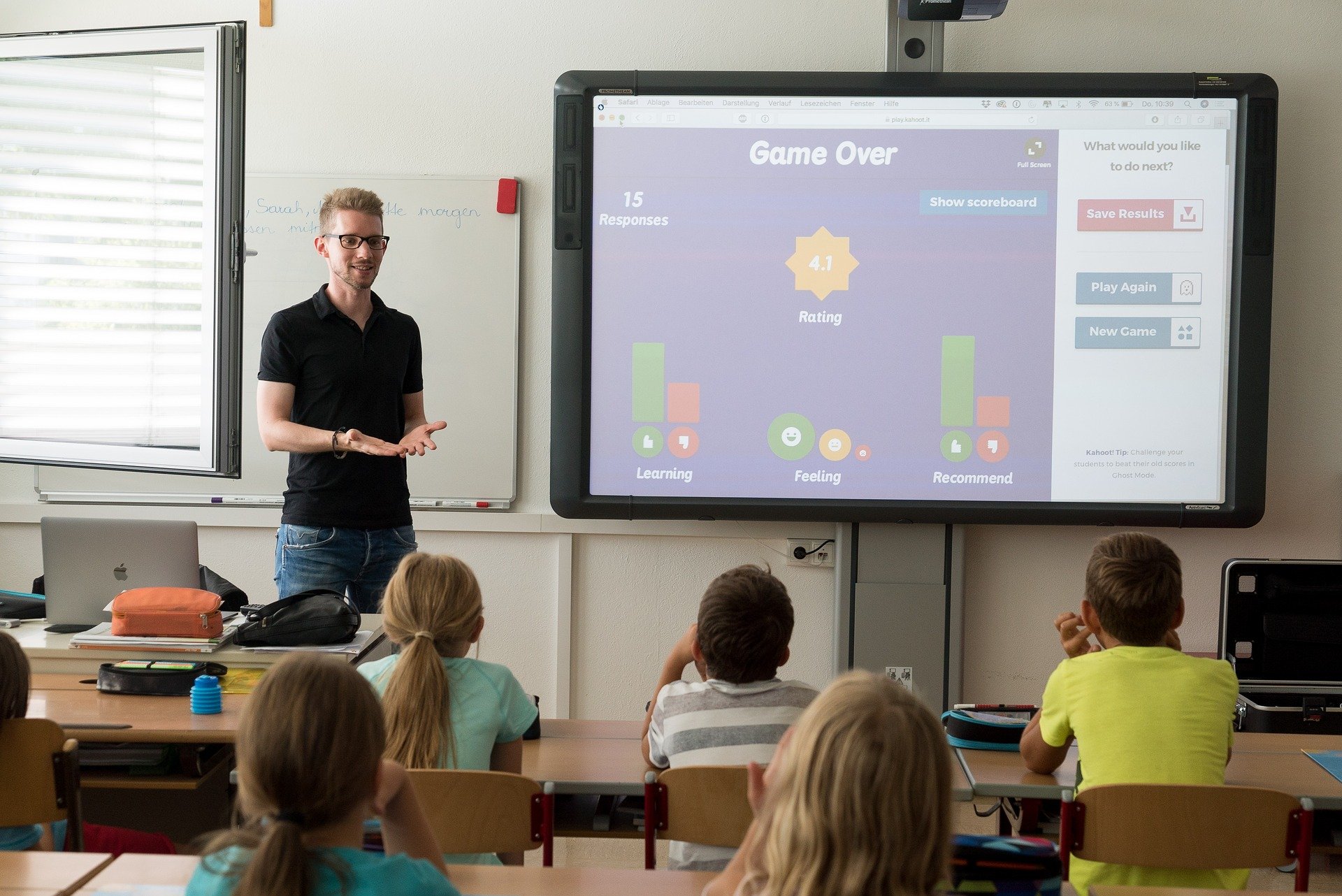 Nauczyciel stoi w klasie przed uczniami, za nim widoczny jest duży ekran na którym wyświetlają się informacje w jezyku angielskim