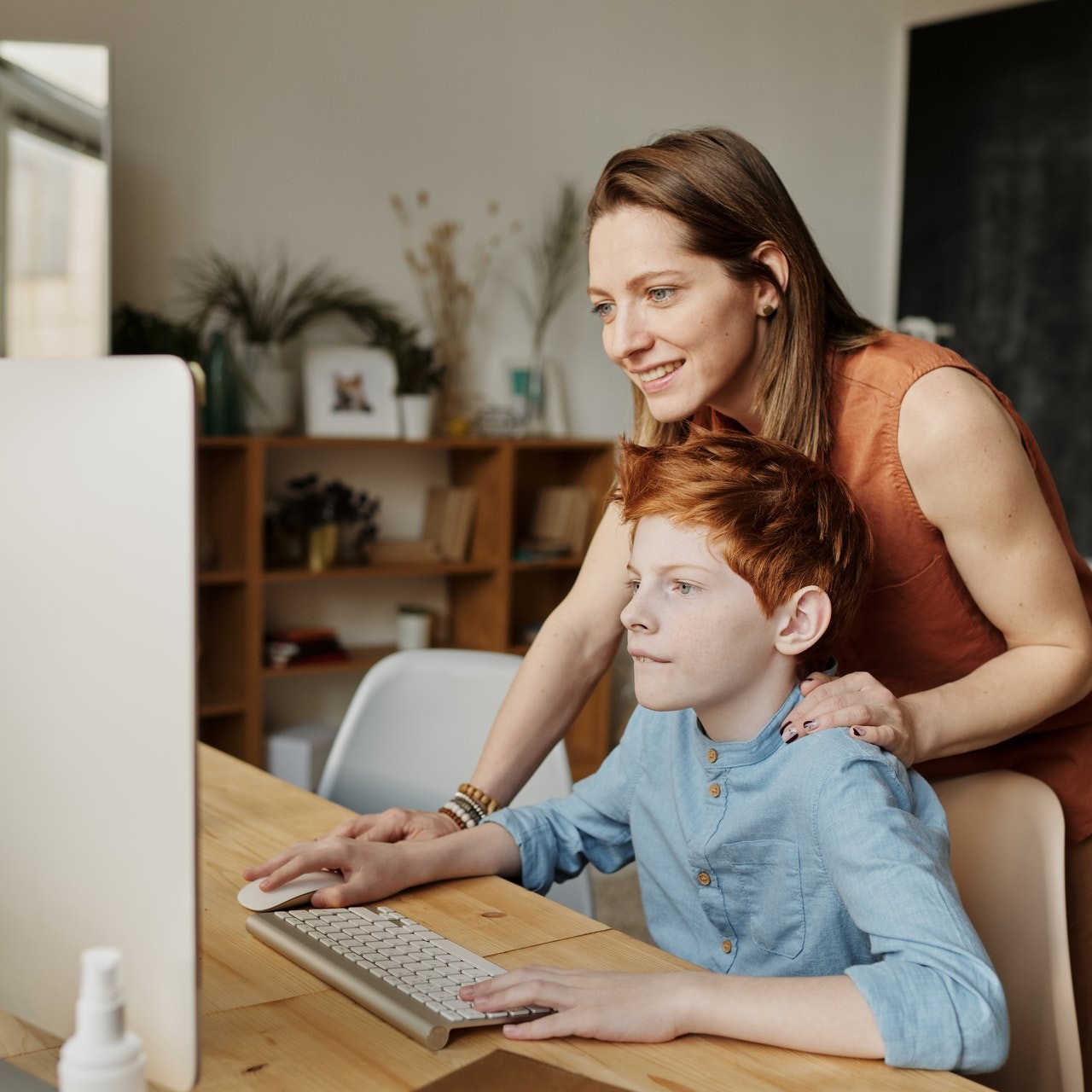 Chłopiec patrzy ze skupieniem w ekran monitora. Za nim stoi kobieta (mama) trzymając jedną dłoń na myszce komputerowej a drugą na ramieniu chłopca.
