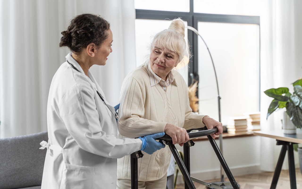 starsza kobieta podpierającą się na chodziku rehabilitacyjnym, obok stoi lekarka